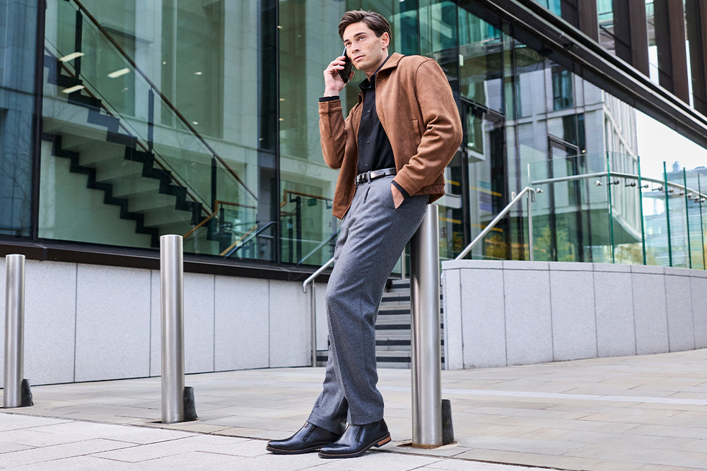 A man on a city pavement leaning against a bollard wearing black leather boots and business wear.