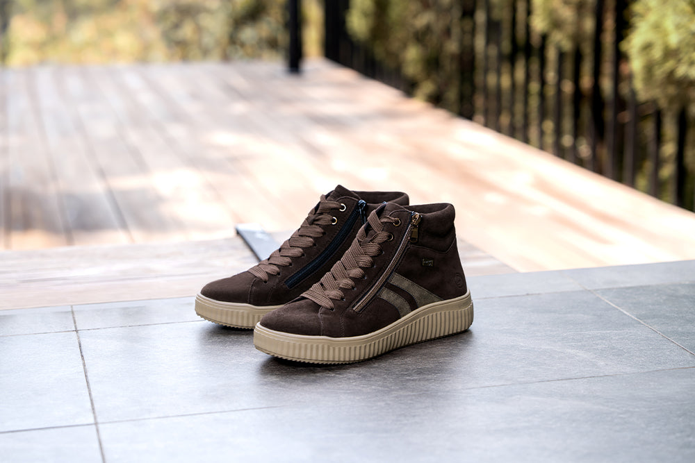 A pair of dark brown high-top trainers with beige soles on a wooden deck with greenery in the background.