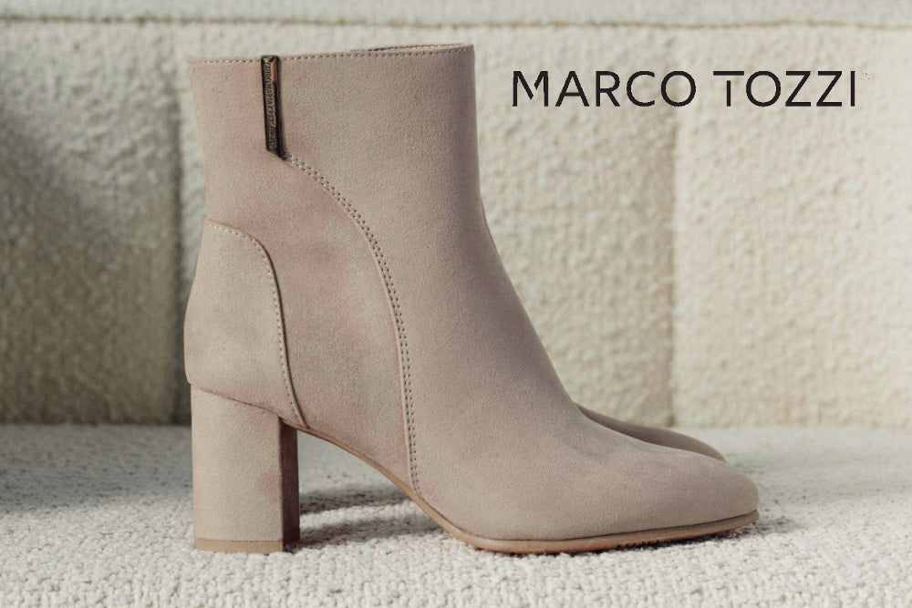 A light taupe suede ankle boot with a block heel and square toe, shown from the side against a textured beige background. The brand name 'MARCO TOZZI' is printed in black text in the upper right corner.